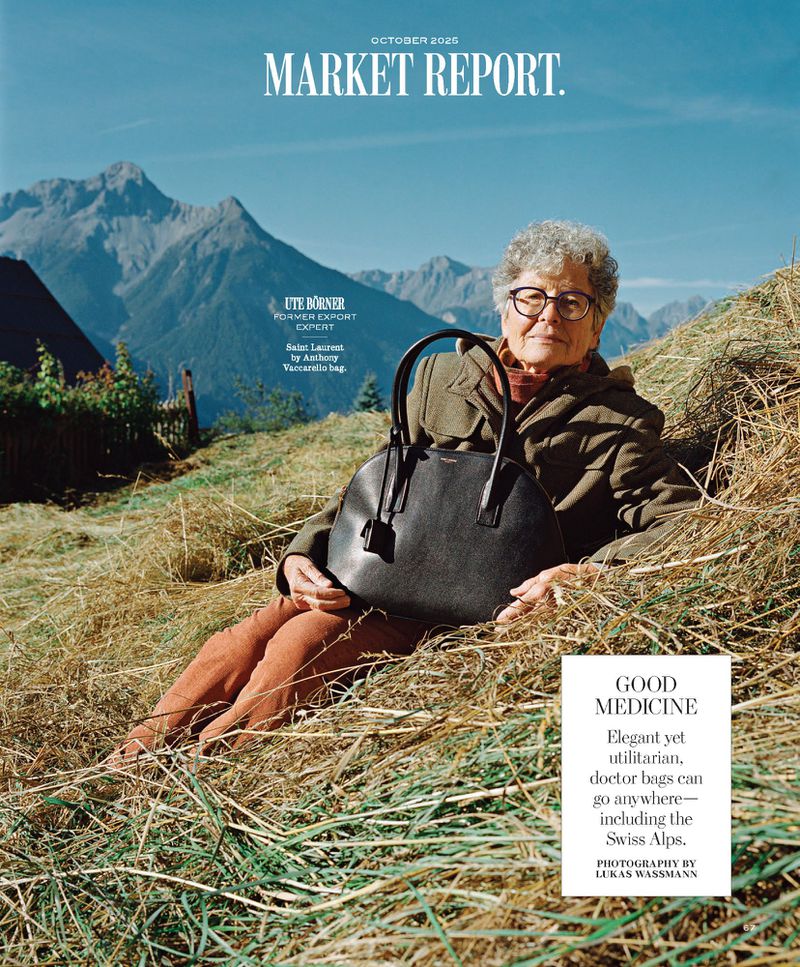 Older woman poses with a black leather handbag in a hay field with Swiss Alps mountains in the background under a blue sky.