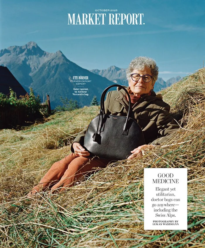 Elderly woman in eyeglasses and an olive jacket reclining in a hayfield with a black leather handbag against a mountain backdrop.