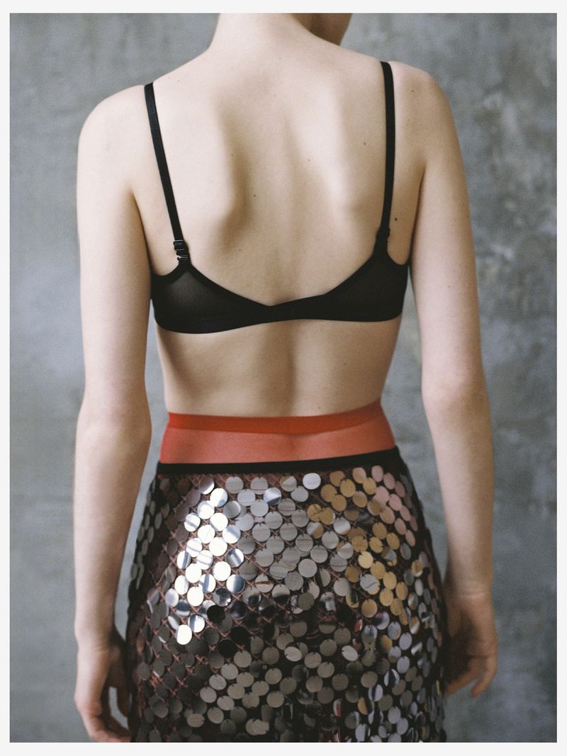 Back view of a woman in a bra and a skirt with reflective circles against a textured gray background in a studio setting.