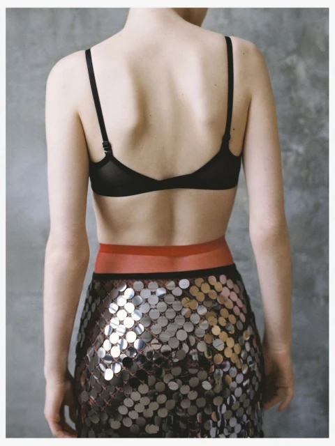 Back view of a woman in a bra and a skirt with reflective circles against a textured gray background in a studio setting.