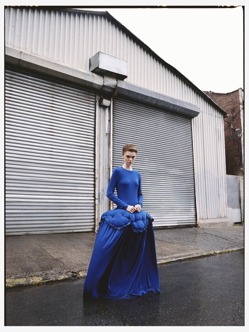 An adult female model poses in a long-sleeved blue dress in an urban setting, featuring a metal warehouse facade and wet pavement.