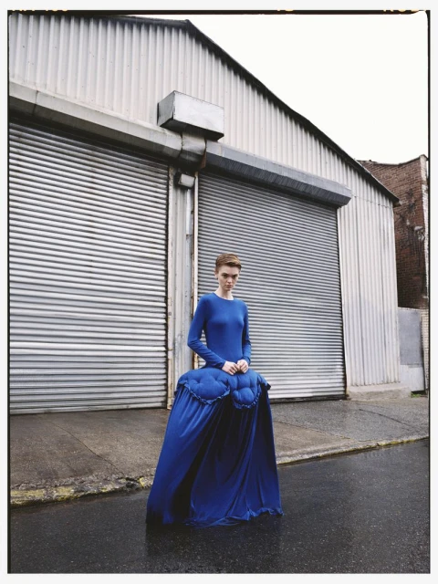 An adult female model poses in a long-sleeved blue dress in an urban setting, featuring a metal warehouse facade and wet pavement.
