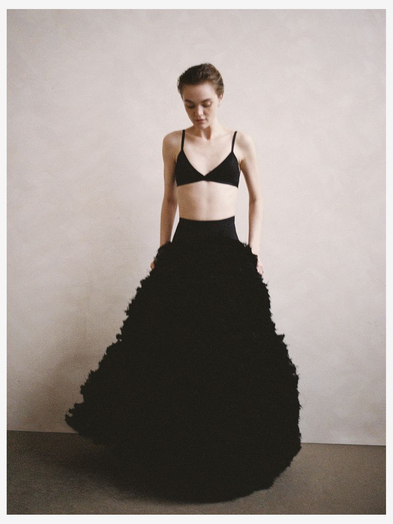 A woman poses indoors in a black bra and a voluminous, ruffled black skirt against a neutral background, creating a fashion-editorial look.