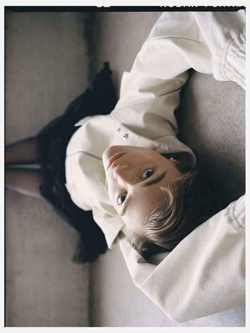 Eye-level portrait of a young adult woman lying on the floor in a cream jacket, black skirt, and jewelry, looking up at the camera.