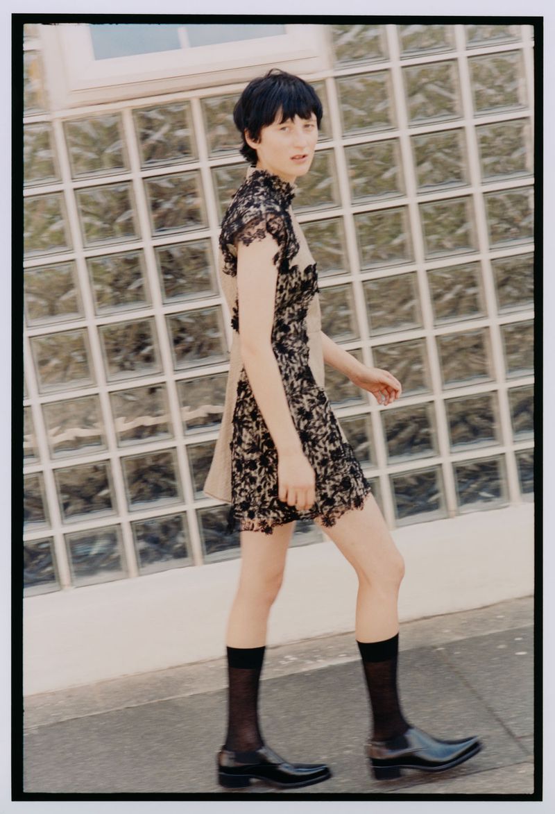 A slender adult fashion model poses in a lace dress with black sheer socks and black patent leather shoes, walking in front of a glass-block wall.