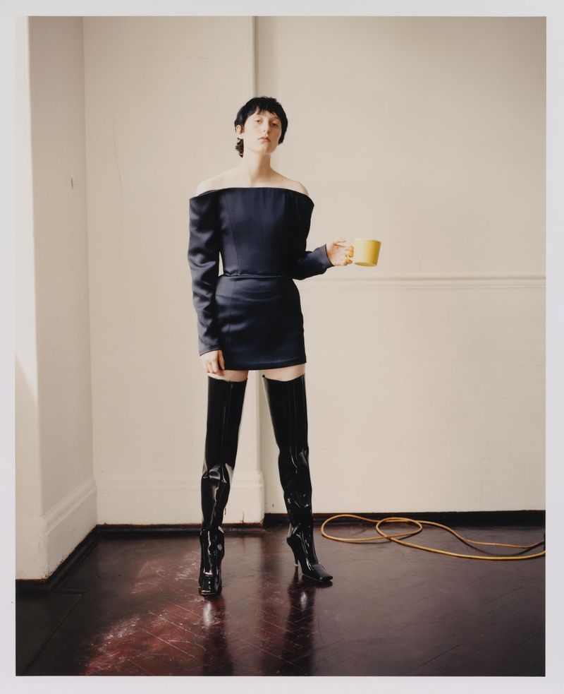 A woman in a black dress and thigh-high boots stands in a room, holding a yellow mug, with a minimal background and studio lighting.