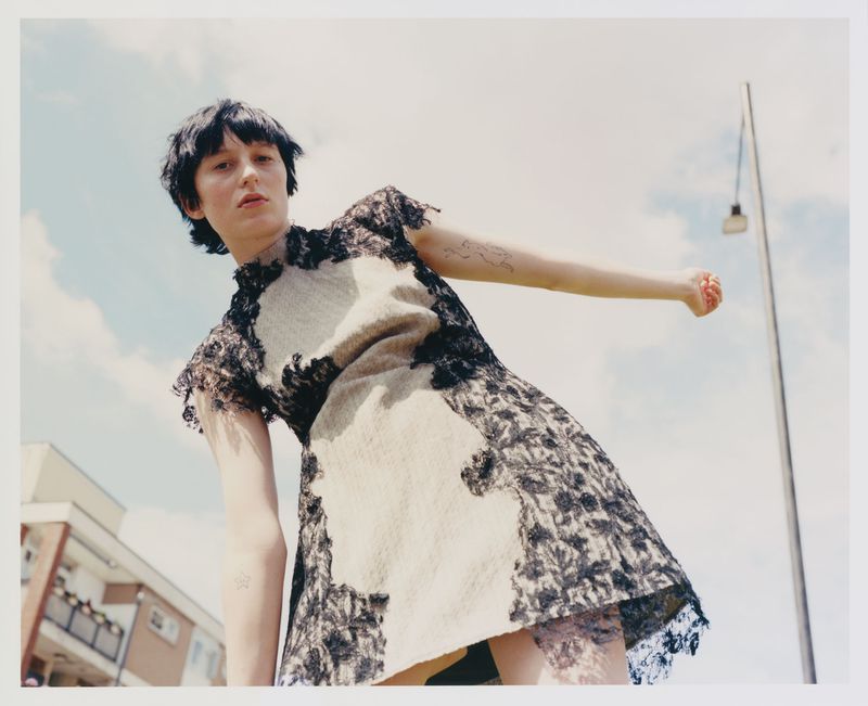 Woman poses in a short, cream-colored dress trimmed with black lace against a blue sky and city buildings in the background. 