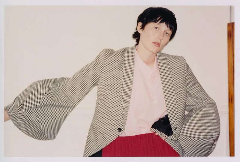Adult female poses indoors wearing a pink shirt, red skirt, and a patterned blazer with oversized sleeves against a neutral wall.