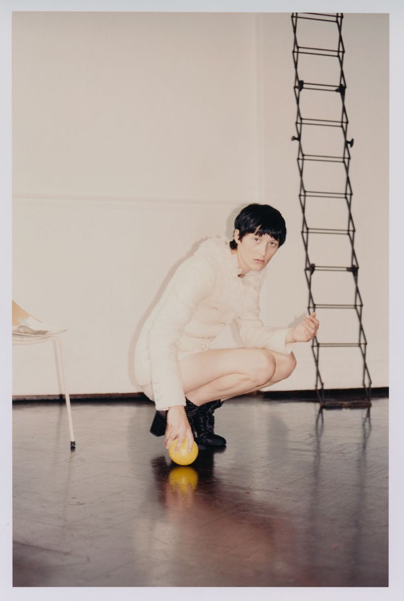 Adult crouches indoors, wearing a light-colored long-sleeved outfit with dark boots, holding a yellow ball near a white chair and black ladder.