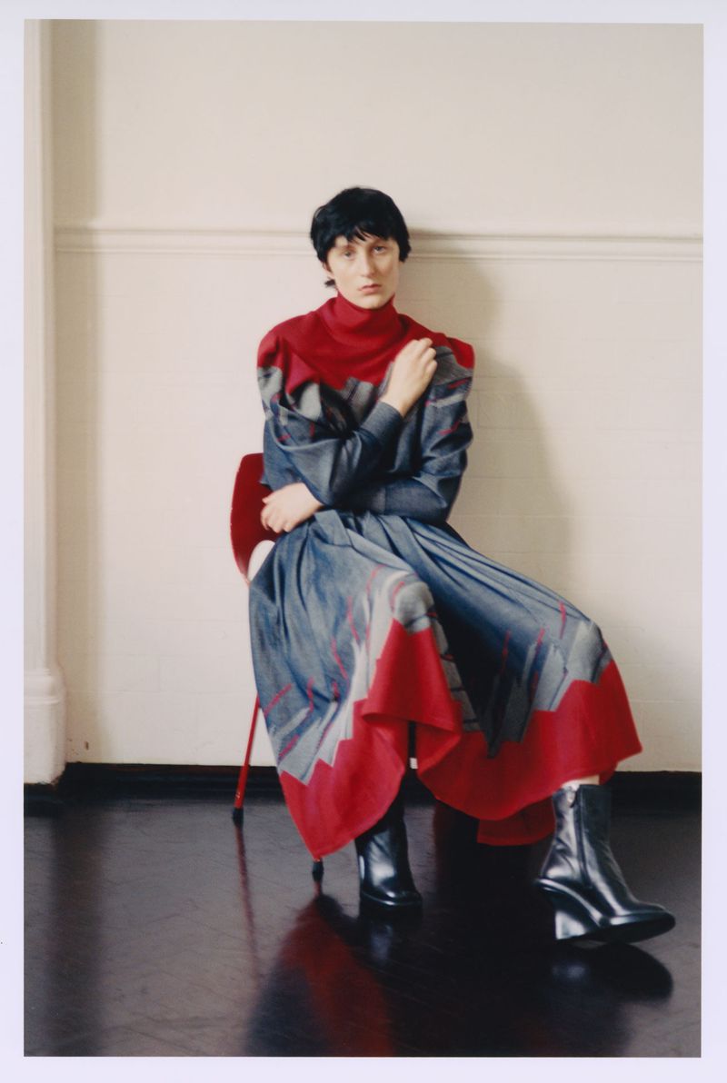 Model with short dark hair seated on a red chair wearing a red and grey patterned pleated dress and black wedge boots.