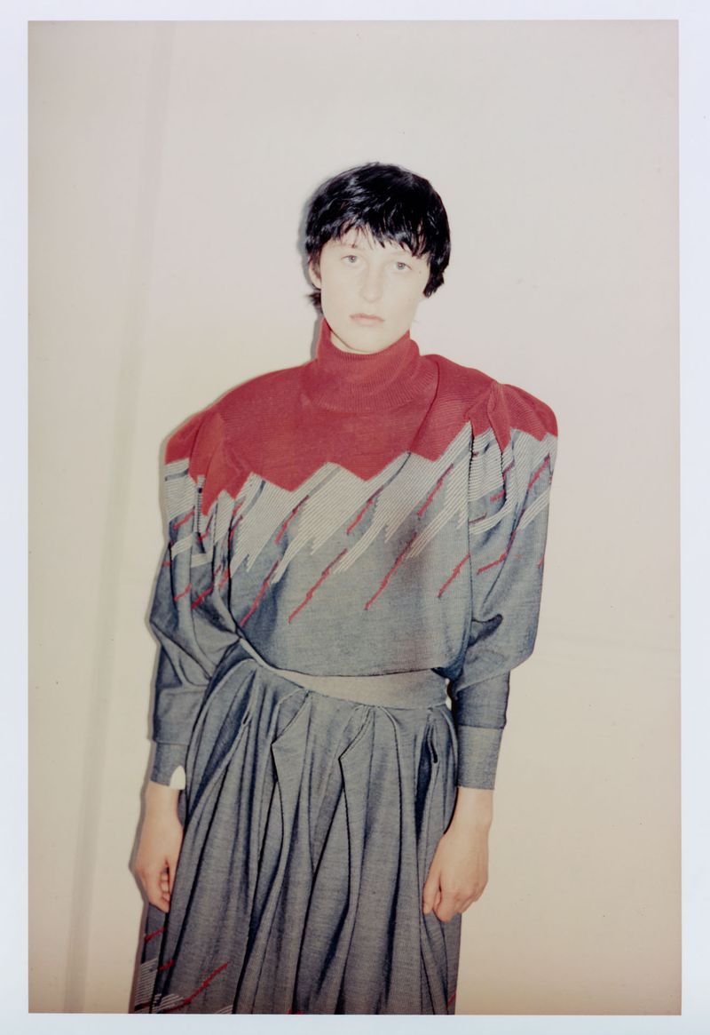 Model Onyx B wearing a high-neck grey and red structured top with a matching pleated skirt against a plain studio background.