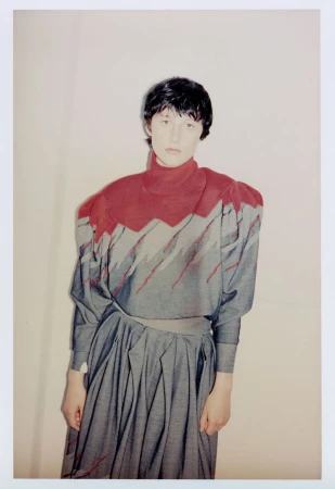 Model Onyx B wearing a high-neck grey and red structured top with a matching pleated skirt against a plain studio background.