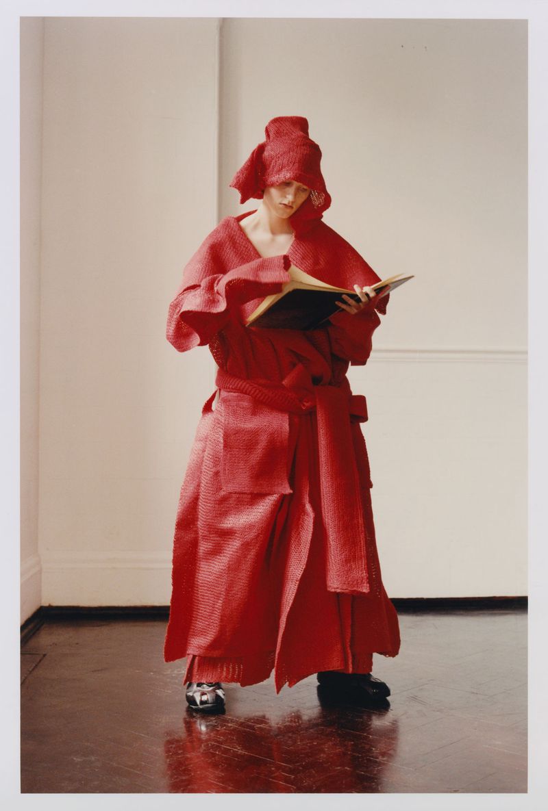 Model in textured red coat and headwear reading a book against a plain interior background
