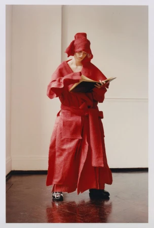 Model in textured red coat and headwear reading a book against a plain interior background