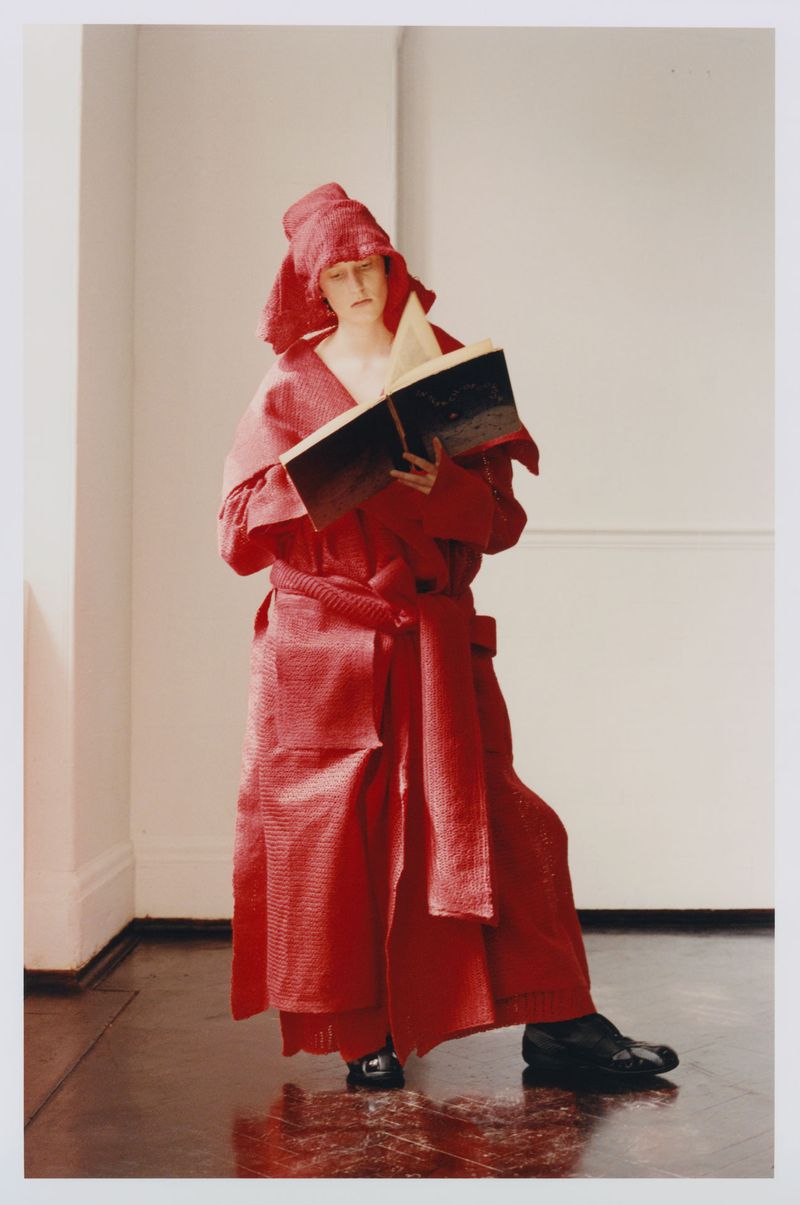 Woman in theatrical red costume and headpiece stands indoors reading a large book with dark cover. Indoor setting with shiny dark floor.