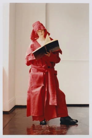 Woman in theatrical red costume and headpiece stands indoors reading a large book with dark cover. Indoor setting with shiny dark floor.