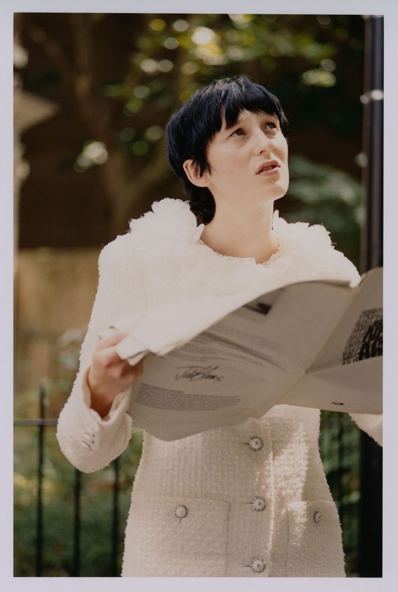 Woman in white jacket holds newspaper outdoors in daylight with trees in background, looking up, fashion editorial shot.