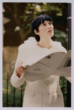 Woman in white jacket holds newspaper outdoors in daylight with trees in background, looking up, fashion editorial shot. 