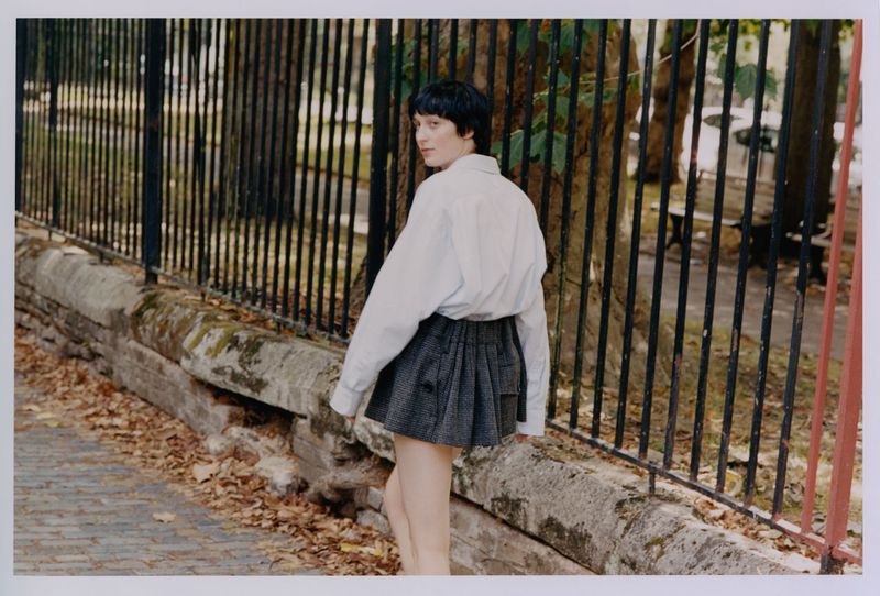 Adult female in oversized shirt and skirt walking past fence and foliage in outdoor park setting, glancing back over her shoulder.