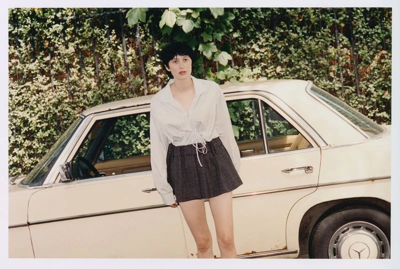 A young adult woman poses next to a vintage beige car with a green leafy background. She wears a button-up shirt and gray plaid skirt.