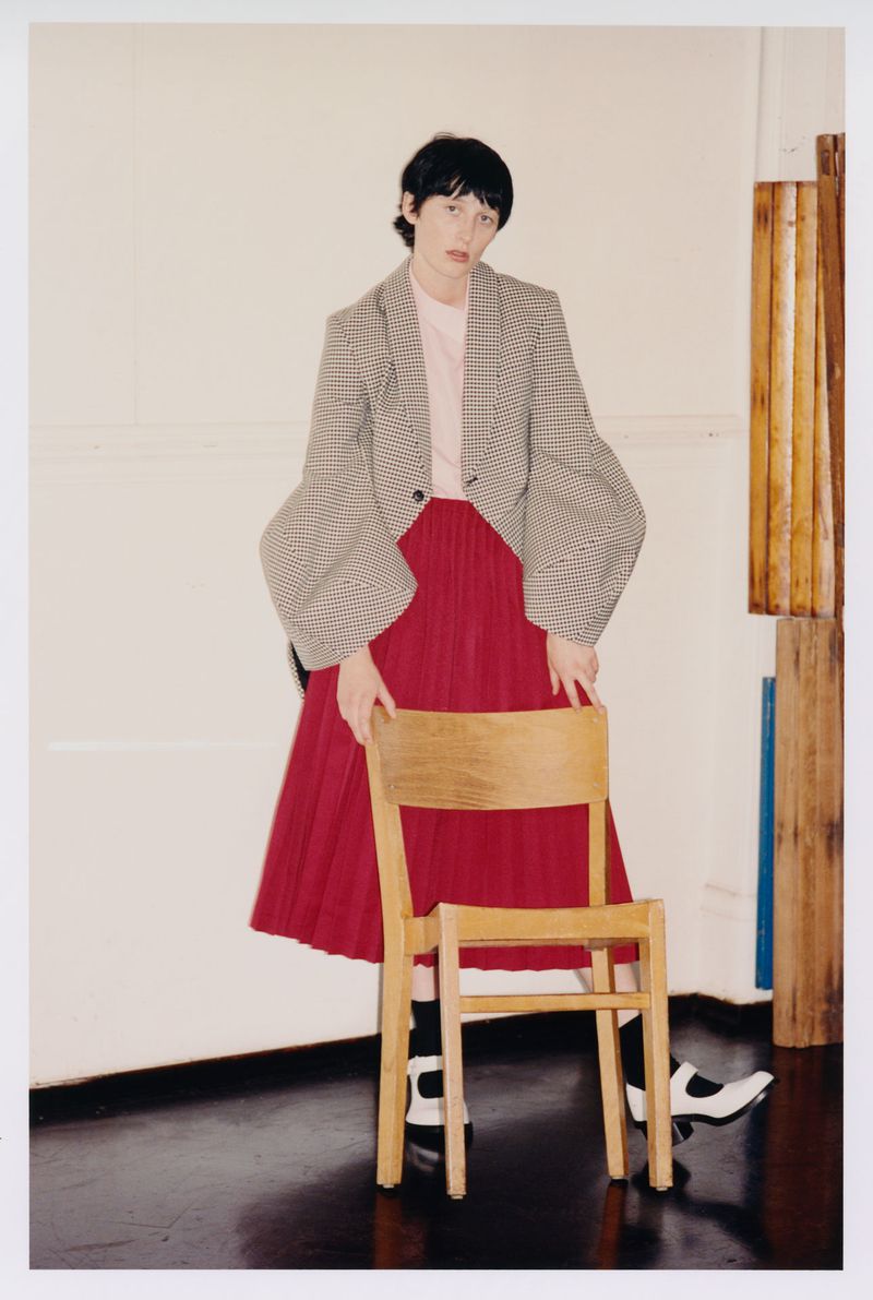 Full shot of a woman in a grey blazer and red skirt holding a wooden chair in a bright indoor space, fashion editorial style.