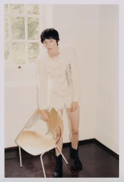 Model in cream jacket leaning on a wooden chair near a window for Lula Japan