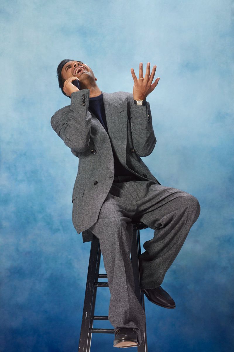 Man in gray suit sitting on a ladder while talking on a cell phone against a blue background in a studio setting with bright lighting.