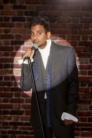 Man in suit holding microphone on stage against brick wall with spotlight. Comedian or speaker performing in front of an audience.
