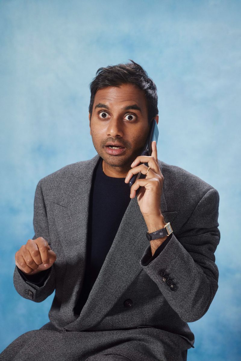Adult male in a gray suit and dark shirt talks on a smartphone. He has a surprised expression, indoors against a mottled blue background.