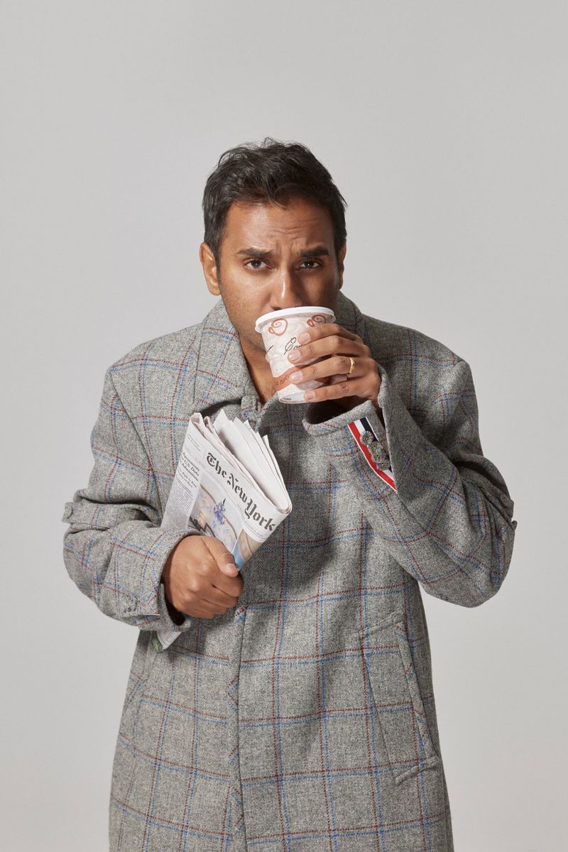 Adult man in a grey coat drinks from a paper cup while holding the New York Times newspaper on a light grey background.