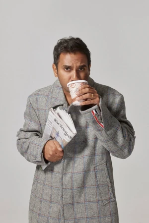 Adult man in a grey coat drinks from a paper cup while holding the New York Times newspaper on a light grey background.