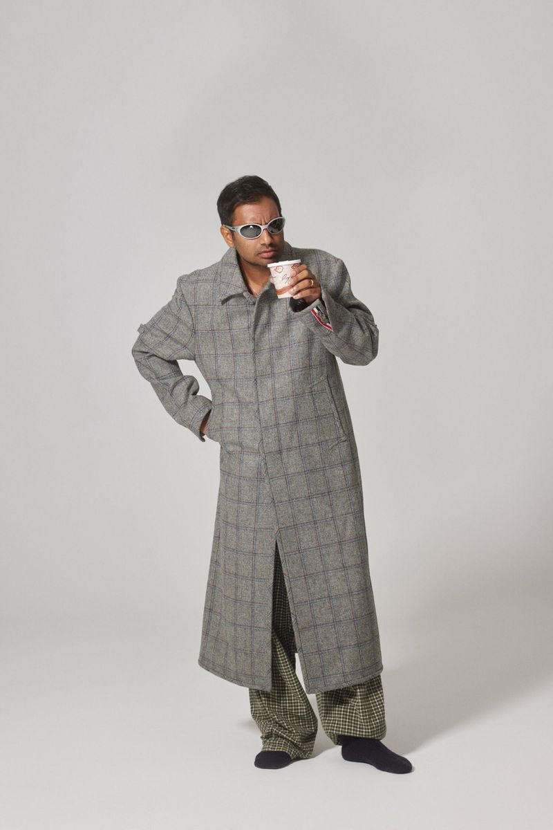 Man in long grey coat, patterned pants, sunglasses, and socks, holding a coffee cup in a white studio environment with soft lighting.