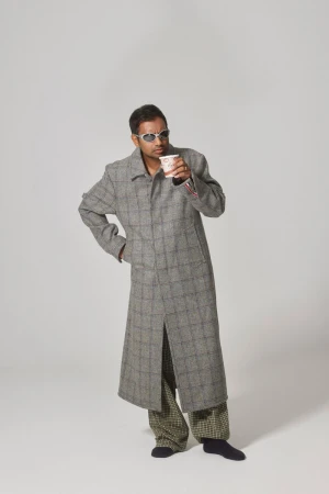 Man in long grey coat, patterned pants, sunglasses, and socks, holding a coffee cup in a white studio environment with soft lighting.