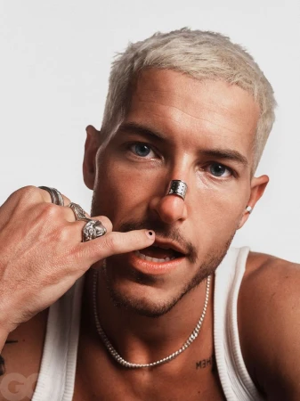 Close-up portrait of a young adult male with a nose ring, a silver chain necklace, and multiple rings on his left hand in a studio setting.