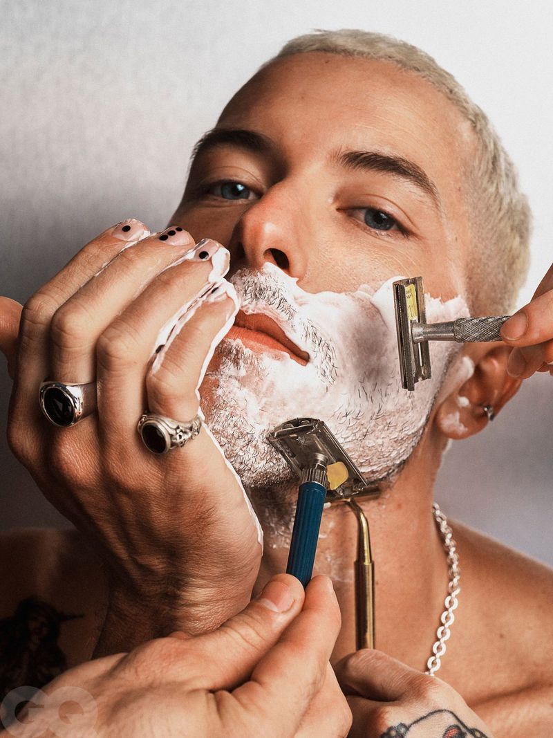 Close-up of a fair-skinned adult man with short bleached hair and multiple hands shaving him with straight razors against a white wall.