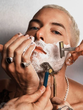 Close-up of a fair-skinned adult man with short bleached hair and multiple hands shaving him with straight razors against a white wall.