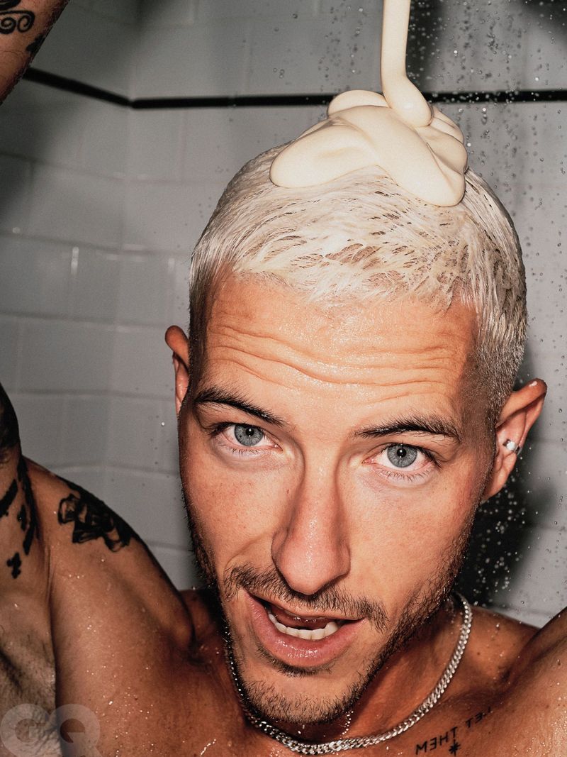 Close-up studio shot of a shirtless adult male with bleached hair having shampoo poured on his head, wearing a chain necklace.