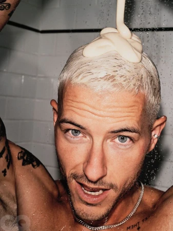 Close-up studio shot of a shirtless adult male with bleached hair having shampoo poured on his head, wearing a chain necklace.