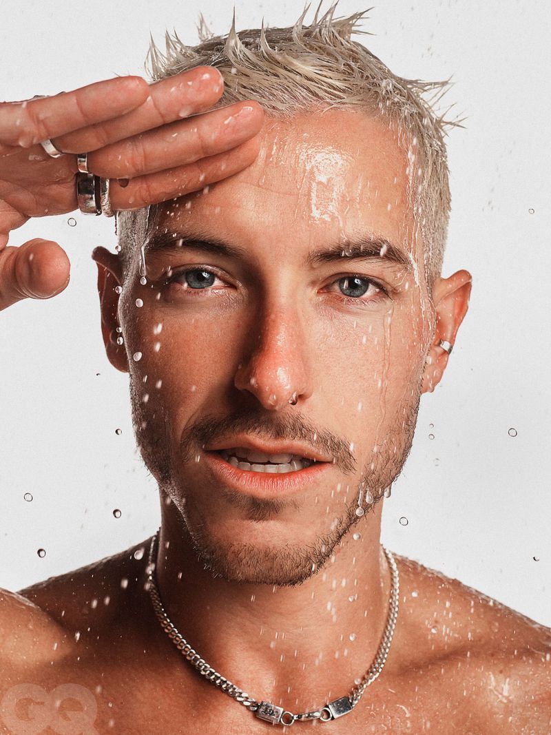 Close-up portrait of a fair-skinned male model with platinum blond hair and light facial hair, posing with water droplets on his face.