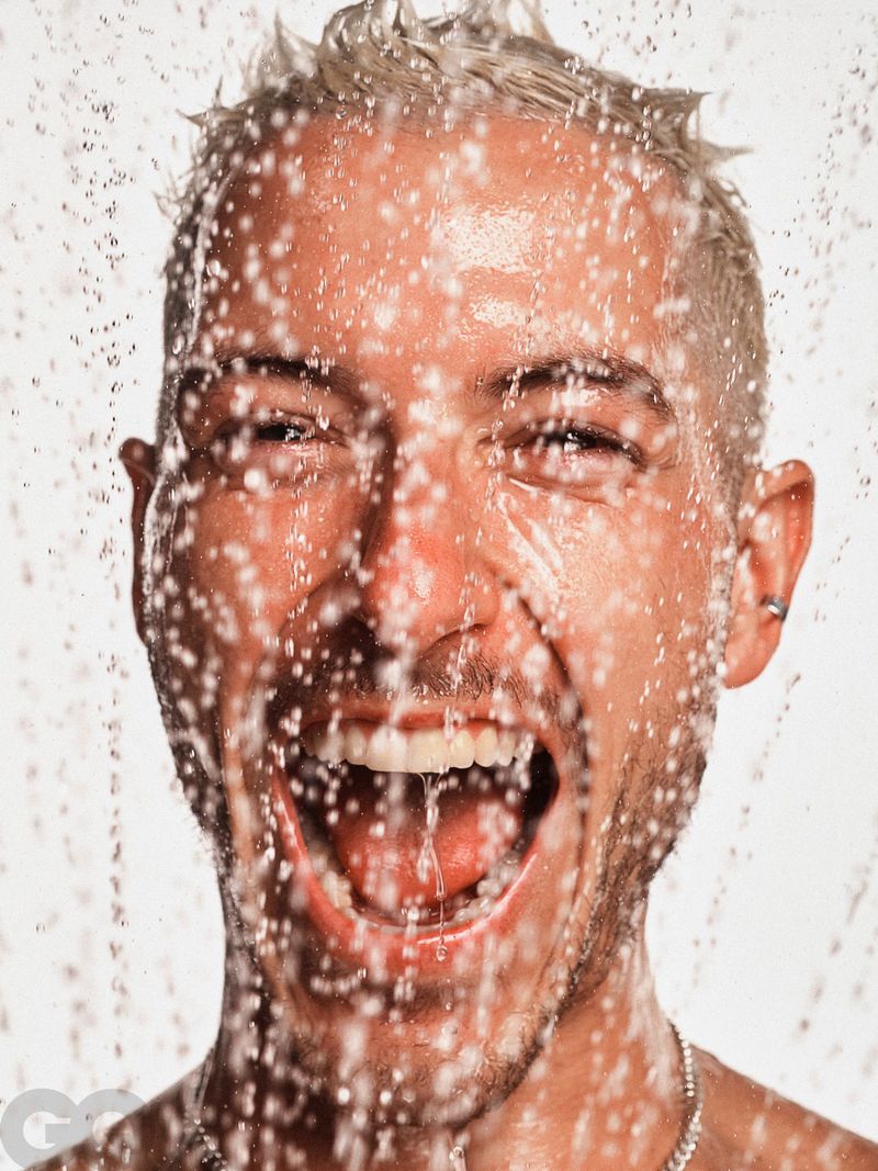Close-up portrait of Taz Skylar shouting with mouth wide open under splashing water against a white background.