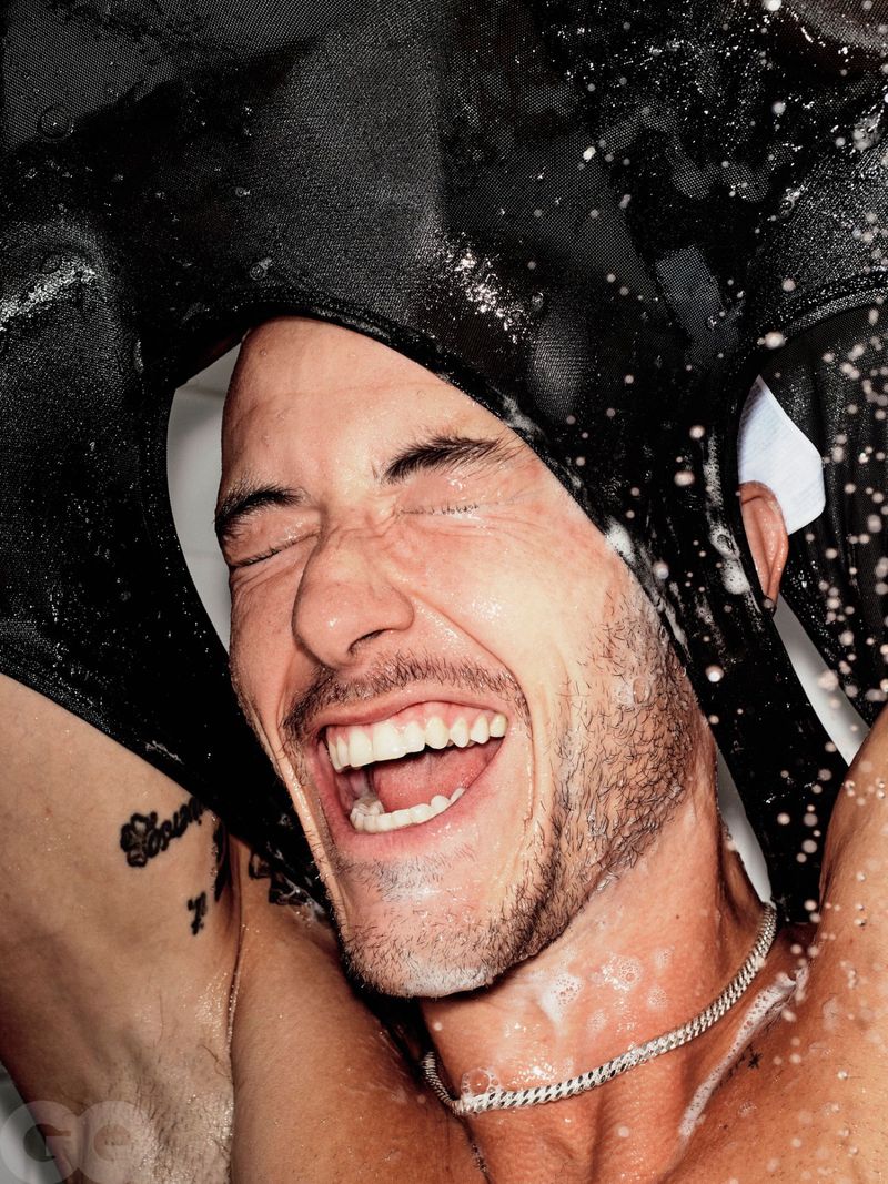 A joyful man with dark hair laughs, water droplets on his face, a black garment draped over his head, set against a neutral background.