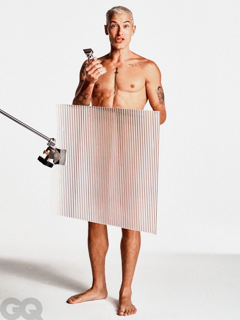 A topless adult male, fair skin, silver hair, holding an electric razor and a ribbed panel in a light gray studio setting.