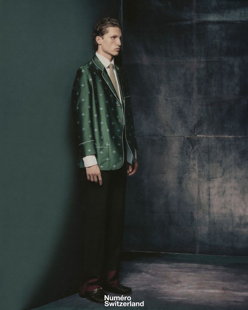 Young man in green patterned blazer with white shirt and tie poses against a dark wall in a fashion editorial studio shot.