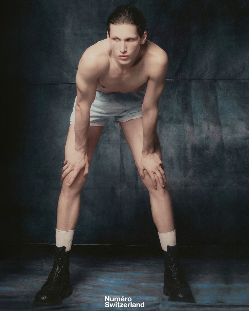 Shirtless adult male fashion model with hands on knees wearing boxer shorts, socks, and boots in a studio with a dark background.