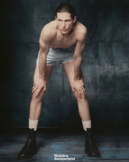 Shirtless adult male fashion model with hands on knees wearing boxer shorts, socks, and boots in a studio with a dark background.