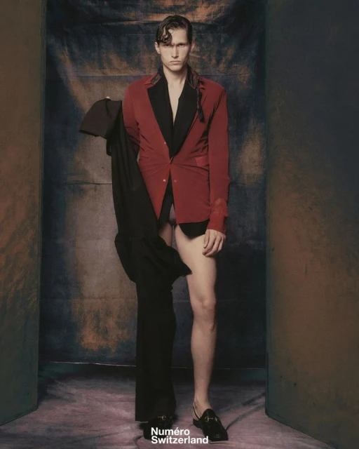 Editorial fashion portrait of a male model in a red blazer and black loafers holding a suit jacket against a studio backdrop.