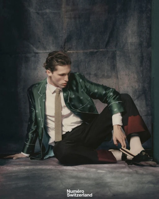A fashionable adult male poses indoors on the floor in a green patterned jacket, white shirt, tie, and black pants, shot in portrait orientation.