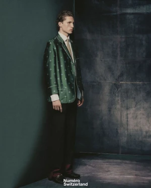 Full-length portrait of a male model in a green patterned blazer standing against a dark wall for Numéro Switzerland.