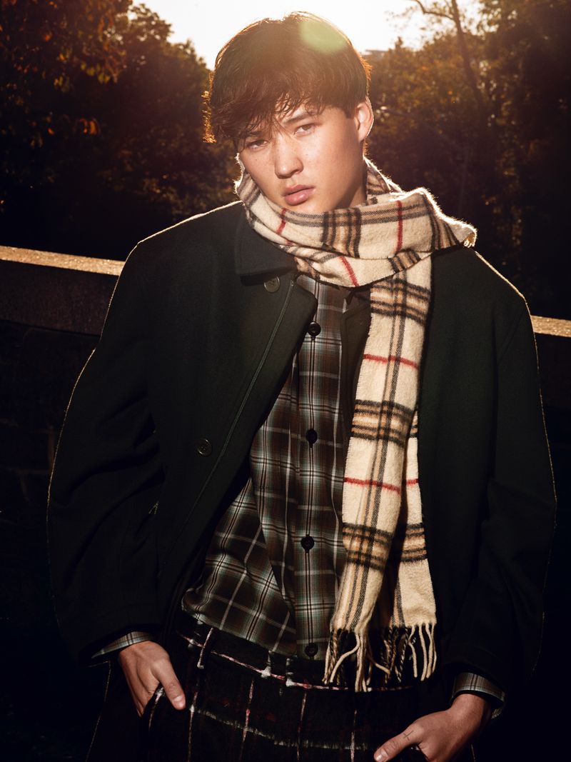 Portrait of a young adult man wearing a plaid scarf and jacket outdoors, standing in front of trees with soft, golden sunlight.