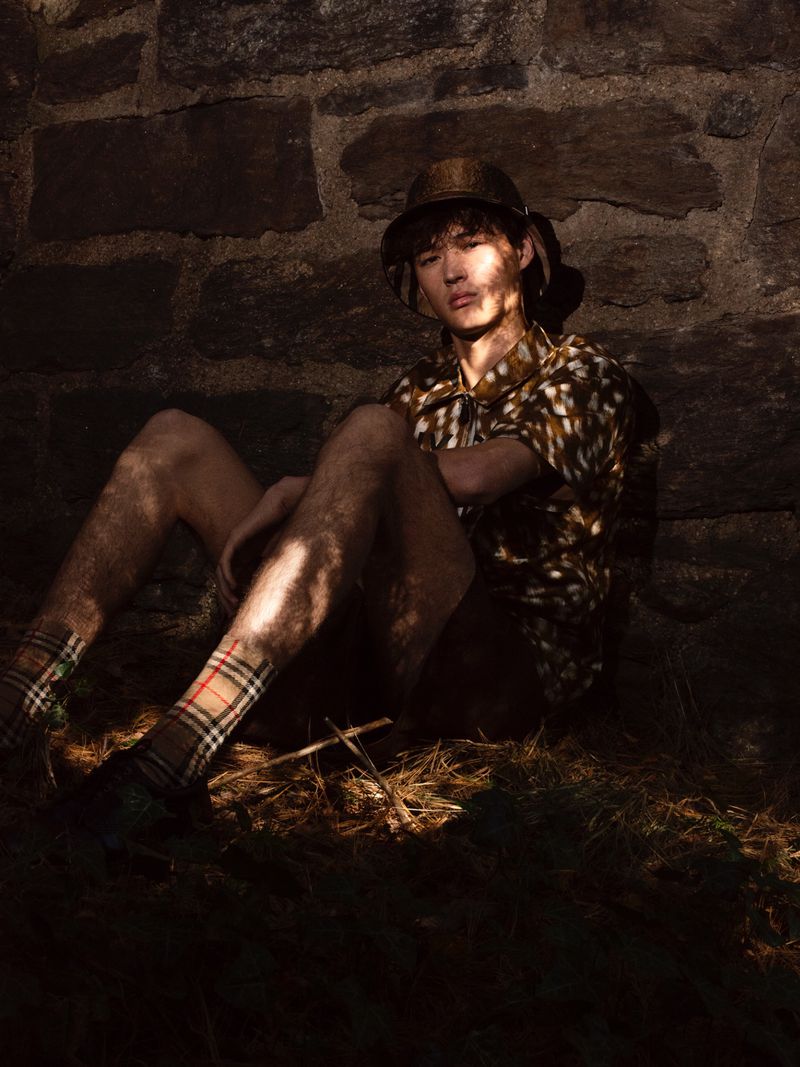 Asian male model in printed shirt and bucket hat poses against a weathered brick wall in shadows. Fashion editorial shot outdoors.